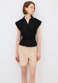 Camisa corta de manga corta negra con cuello, que presenta botones y un lazo en la cintura. Llevada con pantalones cortos beige, de tejido texturizado y diseño a medida.