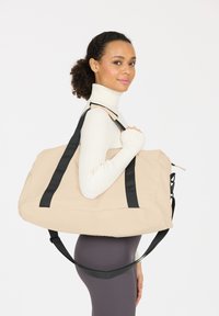 Bolsa de deporte beige con asas negras, fabricada en material suave. Presenta un diseño espacioso con cierre de cremallera y un bolsillo lateral. Ideal para usar en el gimnasio.