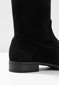 Stivale alla caviglia in suede nero con tacco in gomma nero piatto, caratterizzato da una texture liscia e un design semplice, che mette in evidenza una silhouette leggermente curva.