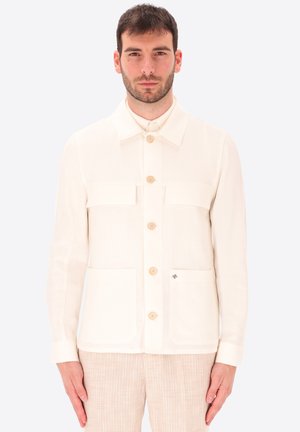 Uomo che indossa una giacca crema con quattro bottoni e tasche applicate, abbinata a pantaloni beige chiaro con texture, in piedi davanti a uno sfondo bianco.