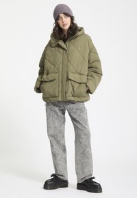 Chaqueta acolchada verde oliva con acolchado en forma de diamante, dos bolsillos frontales y capucha, combinada con jeans texturizados grises y zapatos negros.