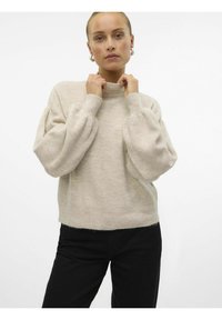 Pull en tricot beige avec un col montant, des manches ballon et une coupe légèrement cropped, associé à un pantalon noir. Texture douce et motif subtil.