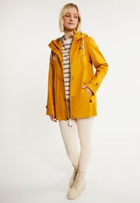 Chaqueta impermeable amarilla con capucha, cintura con cordón y cierres a presión. Combinada con una camiseta a rayas, leggings beige y botas de color claro.