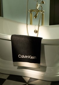 Zwarte handdoeken met een witte rand en de tekst "Calvin Klein" hangen aan een vrijstaande witte badkuip, die gouden hardware en een geometrisch betegelde vloer heeft.