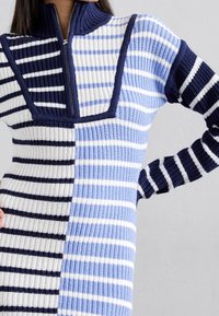 Pull en tricot avec un col montant zippé, présentant un patchwork de rayures bleu clair, bleu marine et blanches, une texture côtelée, et des manches longues.