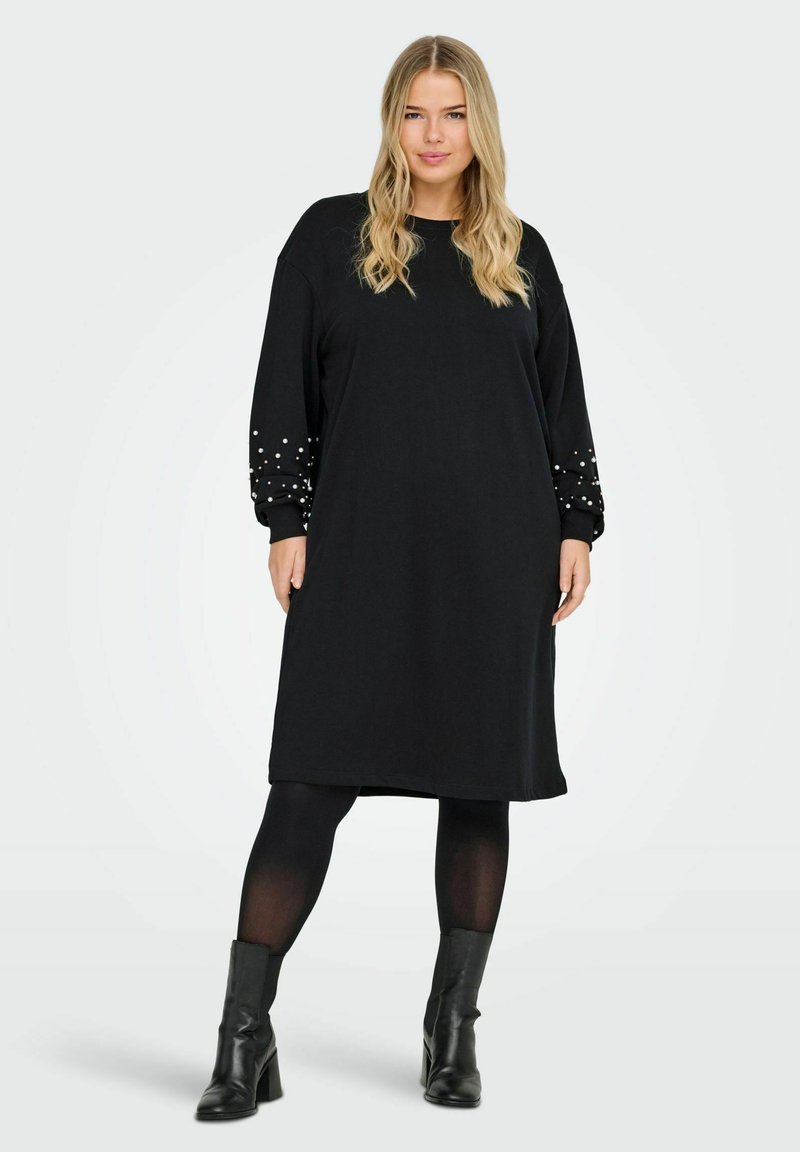 Femme avec de longs cheveux blonds portant une robe noire à manches longues ornée de perles aux manches, des collants noirs et des bottines noires.