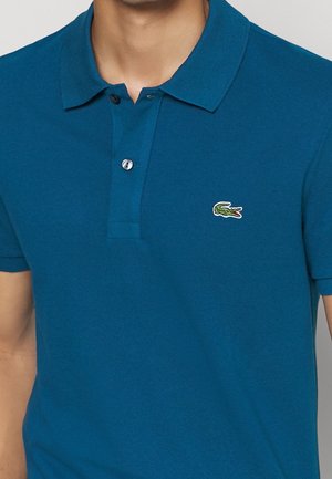 Homme portant un polo bleu sarcelle avec deux boutons et un petit logo crocodile vert sur la poitrine gauche.