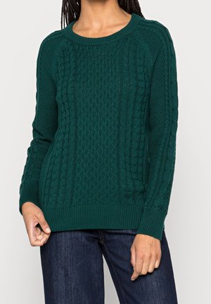 Osoba ubrana w ciemnozielony sweter z długimi rękawami o splocie warkoczowym, w połączeniu z ciemnoniebieskimi jeansami, widziana od szyi do bioder.