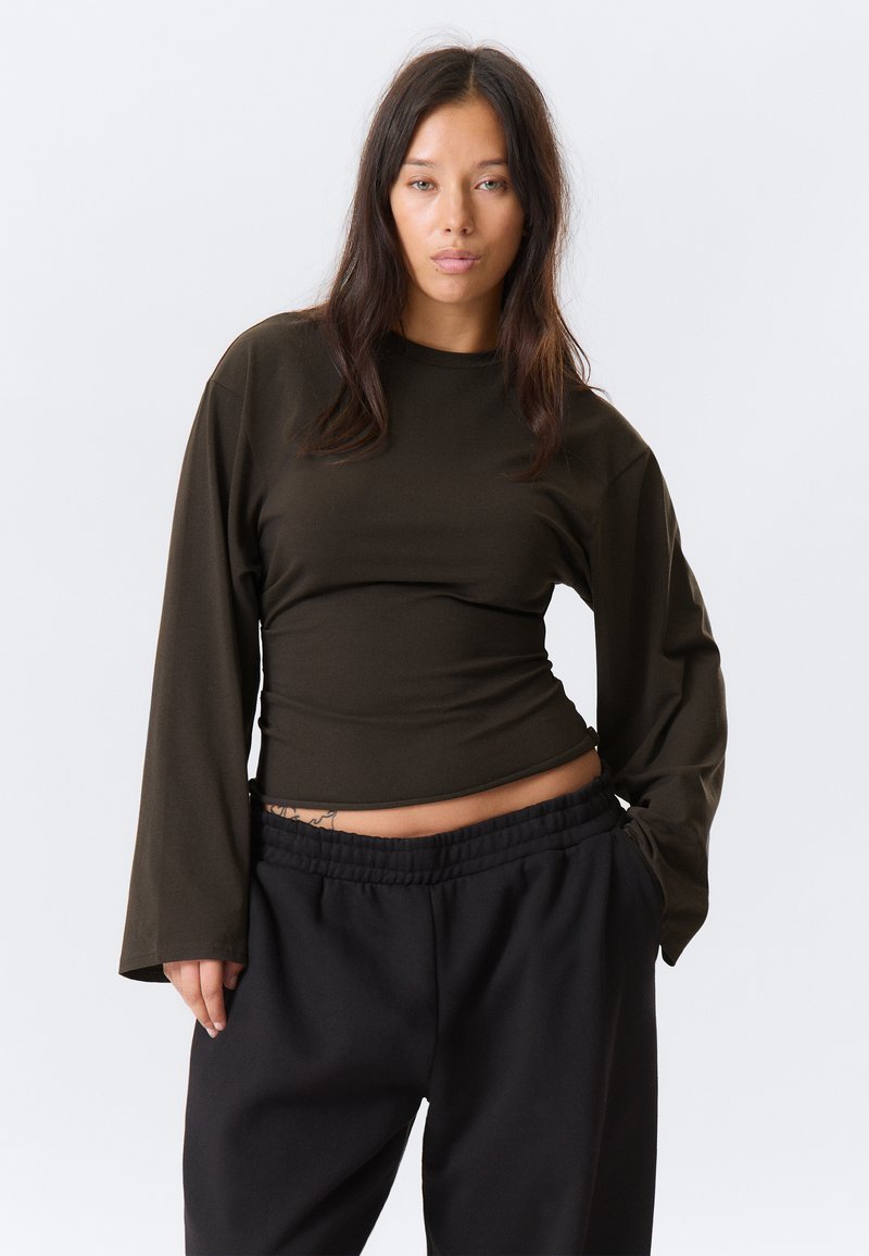 Maglia a maniche lunghe color oliva scuro con bustino aderente e maniche larghe, abbinata a pantaloni neri con vita elastica. Tessuto dalla trama liscia.