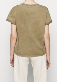 oliwkowa bawełniana koszulka z krótkimi, podwiniętymi rękawami, okrągłym dekoltem i miękką, szczotkowaną teksturą. Dolna część jest lekko zaokrąglona z tyłu.