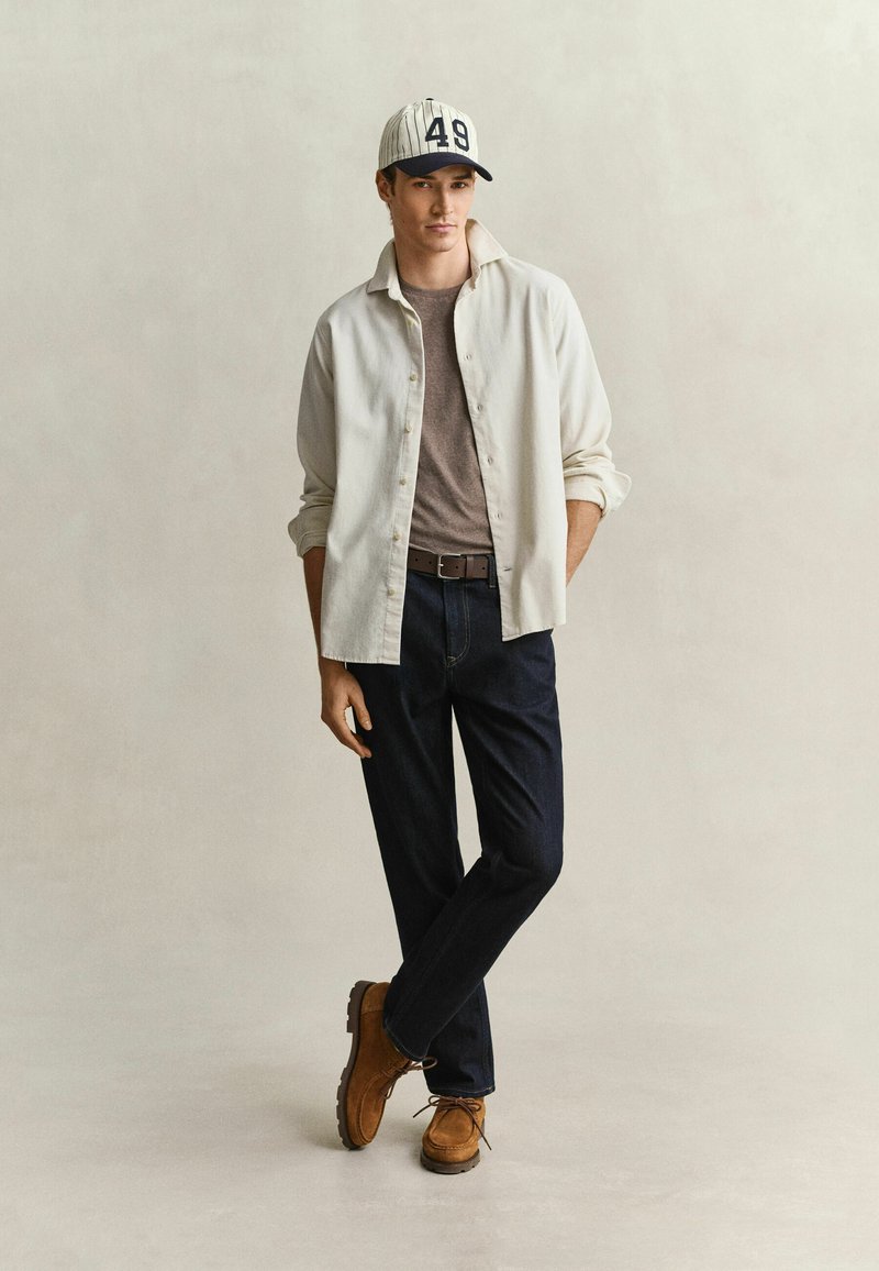 Young man leaning with crossed legs wearing white button shirt, brown t-shirt, dark jeans, brown shoes, and a white baseball cap with number 49.