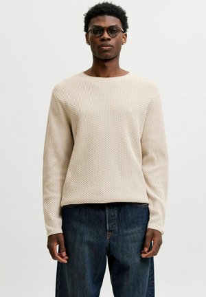 Homme portant des lunettes rondes, un pull beige texturé à manches longues et un jean bleu foncé, debout devant un fond blanc uni.