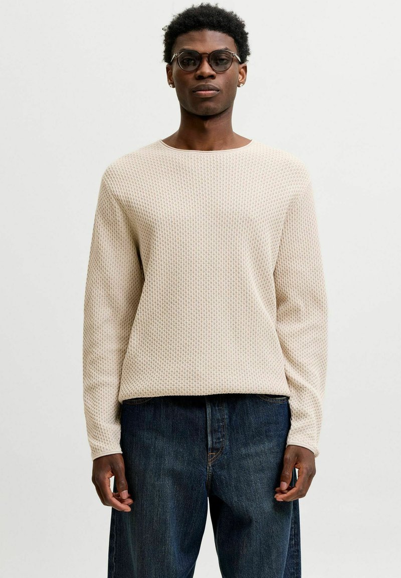 Homme portant des lunettes rondes, un pull beige texturé à manches longues et un jean bleu foncé, debout devant un fond blanc uni.
