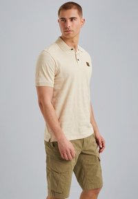 Beige polo-shirt med et struktureret mønster, korte ærmer, tre-knaps placket og et logo-accents på brystet, parret med olivengrønne cargo shorts.