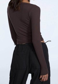 Eine Frau trägt ein enges, braunes Langarmoberteil und lockere schwarze Cargo-Hosen, von hinten vor einem schlichten, hellen Hintergrund gezeigt.