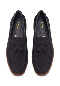 Mocassins en daim bleu marine avec une texture lisse, présentant un gland décoratif et une semelle en caoutchouc marron contrastante. Marque Clarks Signature à l'intérieur.