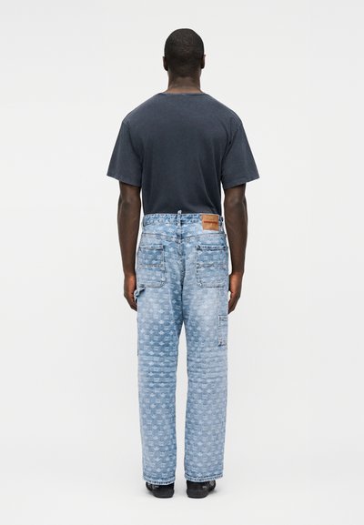 Άνδρας που φοράει ανοιχτό μπλε τζιν με σχέδια και τσέπες-επενδύσεις και μαύρο t-shirt, στέκεται με την πλάτη γυρισμένη σε απλό φόντο.