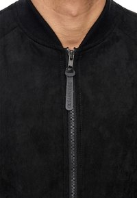 Veste en velours noir avec fermeture éclair à l'avant, col côtelé et fermeture éclair noire contrastante. Fini texturé sans motifs ni accents visibles.