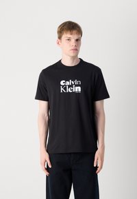 Juodas medvilnės marškinėliai su "Calvin Klein" logotipu baltu šriftu. Apvalus iškirpimas, trumpomis rankovėmis ir įprasta kirpimu. Paprastas dizainas be raštų.