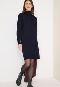 Abito a dolcevita blu navy con maniche lunghe. Realizzato in tessuto testurizzato, presenta una linea dritta. Indossato con stivali da combattimento neri e collant trasparenti.