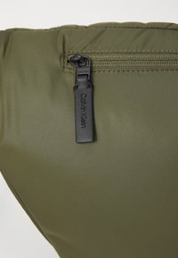 Material acolchado de color verde oliva con una cremallera negra, con una etiqueta de Calvin Klein unida al tirador de la cremallera, indicando la identidad de la marca.