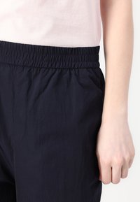 Navy elastisk midjebukser med en rynket tekstur. Nærbilde av en hånd som holder stoffet, som viser den avslappede passformen og materialet.