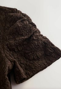 Tissu brun foncé texturé avec un motif de dentelle florale en relief, plié sur une surface blanche.