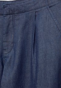 Gros plan sur un pantalon en denim bleu montrant la ceinture, la poche latérale et les coutures sur un tissu uni et texturé. Aucun bouton ni fermeture éclair visible.