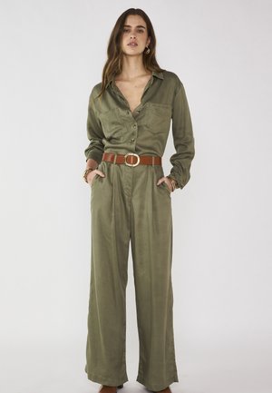 Femme portant une combinaison kaki à manches longues avec jambes larges, ceinture marron, boucles d'oreilles créoles dorées et bracelets, debout avec les mains dans les poches.