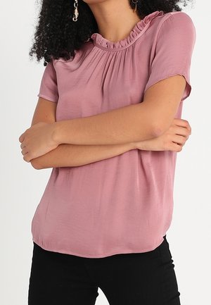 Femme portant un chemisier rose poudré à manches courtes avec un col à volants, bras croisés, et un pantalon noir sur un fond uni.