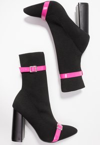 Mustat neulevartalokengät, joissa on terävä nenä, kiiltävät vaaleanpunaiset hihnat ja paksu musta korko, jossa on teksturoitu pinta.