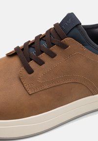 Brown leather sneaker with textured panels, dark brown laces, gray accents, and a white rubber sole. Features a stitched design.