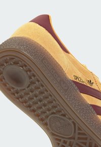 Baskets en daim jaune avec des bandes bordeaux, une semelle en caoutchouc texturée et un logo "adidas" embossé sur le côté. Motif distinctif sur la semelle.