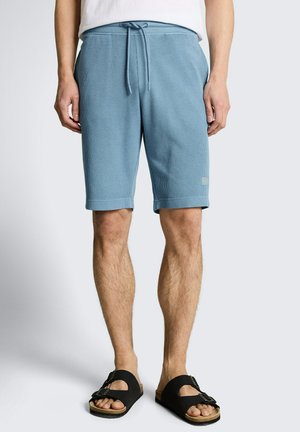 Man die lichtblauwe shorts met trekkoord en zwarte sandalen met gesp draagt, staand tegen een effen lichtgrijze achtergrond.