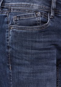 Jeans i denim med en mörkblå tvätt, med en liten framficka, synliga sömmar och en slät yta. Nära beskuren vy.