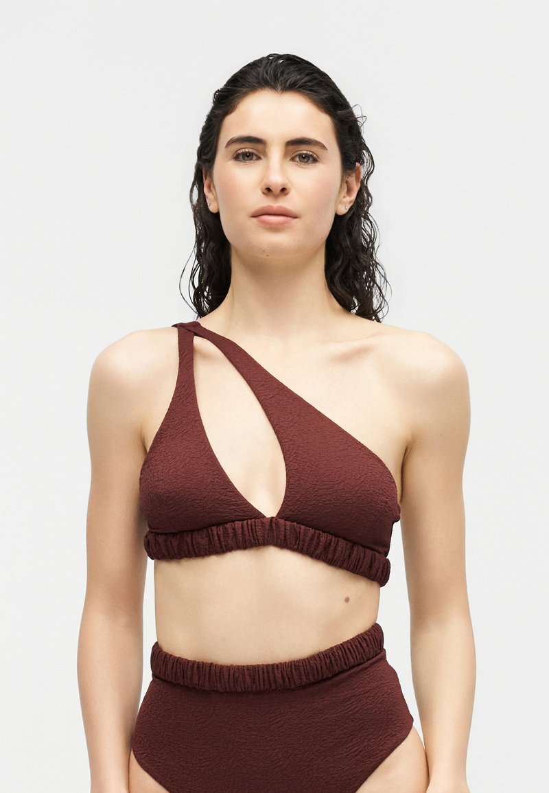 Femme aux cheveux bouclés foncés portant un bikini asymétrique rouge foncé texturé avec un bas taille haute sur un fond blanc uni.