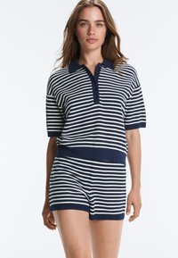 Femme portant un polo à manches courtes rayé horizontalement bleu marine et blanc et un short assorti sur un fond blanc uni.