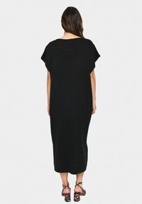 Egy nő laza fekete midi ruhát és leopárdmintás magassarkú szandált visel, háttal állva egy fehér háttér előtt.