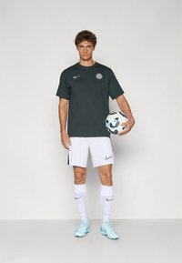 Mladý muž v čiernom športovom tričku, bielych šortkách a modrých kopačkách drží bielo-modrú futbalovú loptu na jednoliatom pozadí.