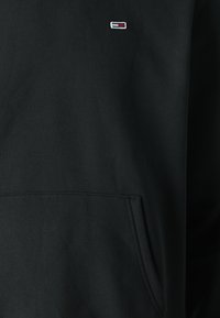 Felpa nera con cappuccio, dotata di una tasca a marsupio frontale e un piccolo logo rettangolare in rosso, bianco e blu cucito sopra la tasca.