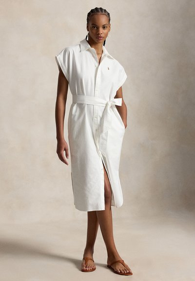 Woman standing wearing a white sleeveless belted midi dress with buttons and tan sandals against neutral background.