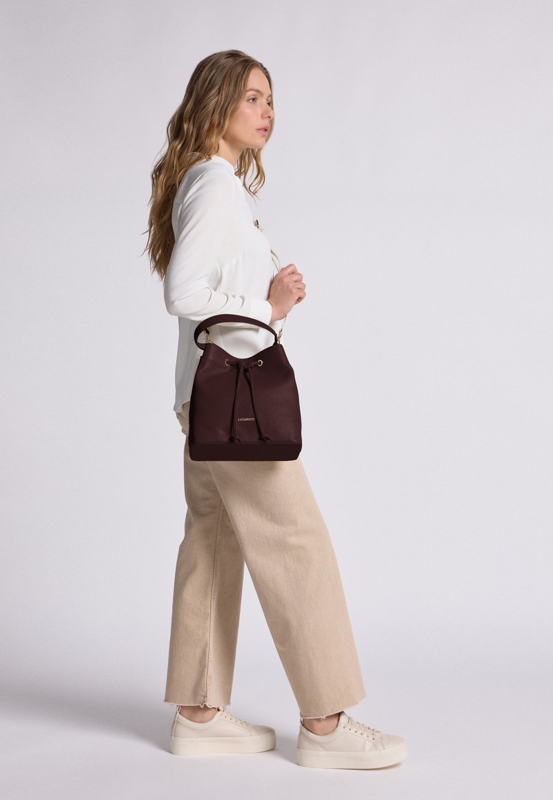 Donna con camicetta bianca e pantaloni beige a palazzo che porta una borsa a secchiello in pelle marrone scuro con dettagli dorati.