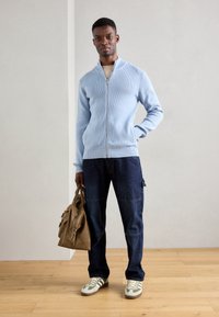 Light blue zip-up sweater with ribbed texture, dark blue cargo pants, and tan bag. Model wears beige and green sneakers. Wooden floor background.
