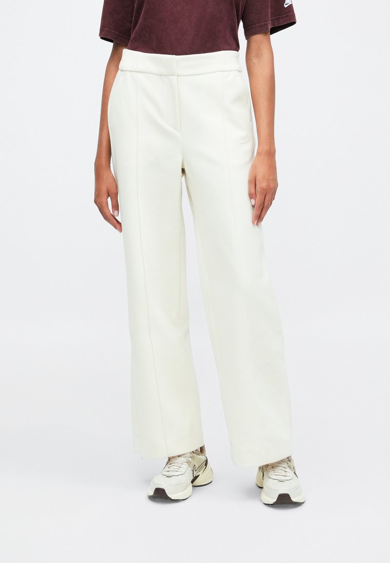 Weiße, weit geschnittene Sweatpants mit glatter Textur und elastischem Bund, kombiniert mit einem figurbetonten weinroten Shirt und neutralen Sneakers.