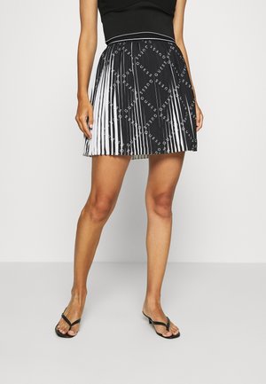 Femme portant une jupe plissée noire et blanche avec des motifs répétés du logo « GUESS », associée à un haut noir et des sandales noires.
