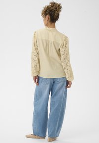 Blouse jaune clair avec manches ballon en dentelle, col à volants et coupe ample, associée à un jean large bleu clair.