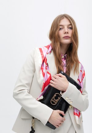 Una mujer con cabello largo lleva un blazer claro y una camisa rosa, sosteniendo un bolso de mano negro con un logo dorado y una bufanda de seda colorida.