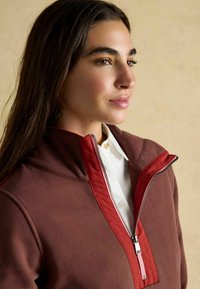 Hnedá bundá na zips s červeným golierom a detailom zipsu, s hladkou textúrou a bielou košeľou pod ňou.