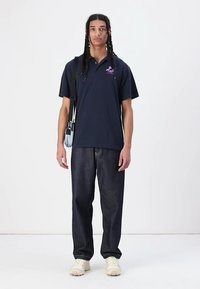 Navy polo shirt with short sleeves, a logo on the chest, paired with dark denim pants and light-colored sneakers. Adjustable bag slung over shoulder.
