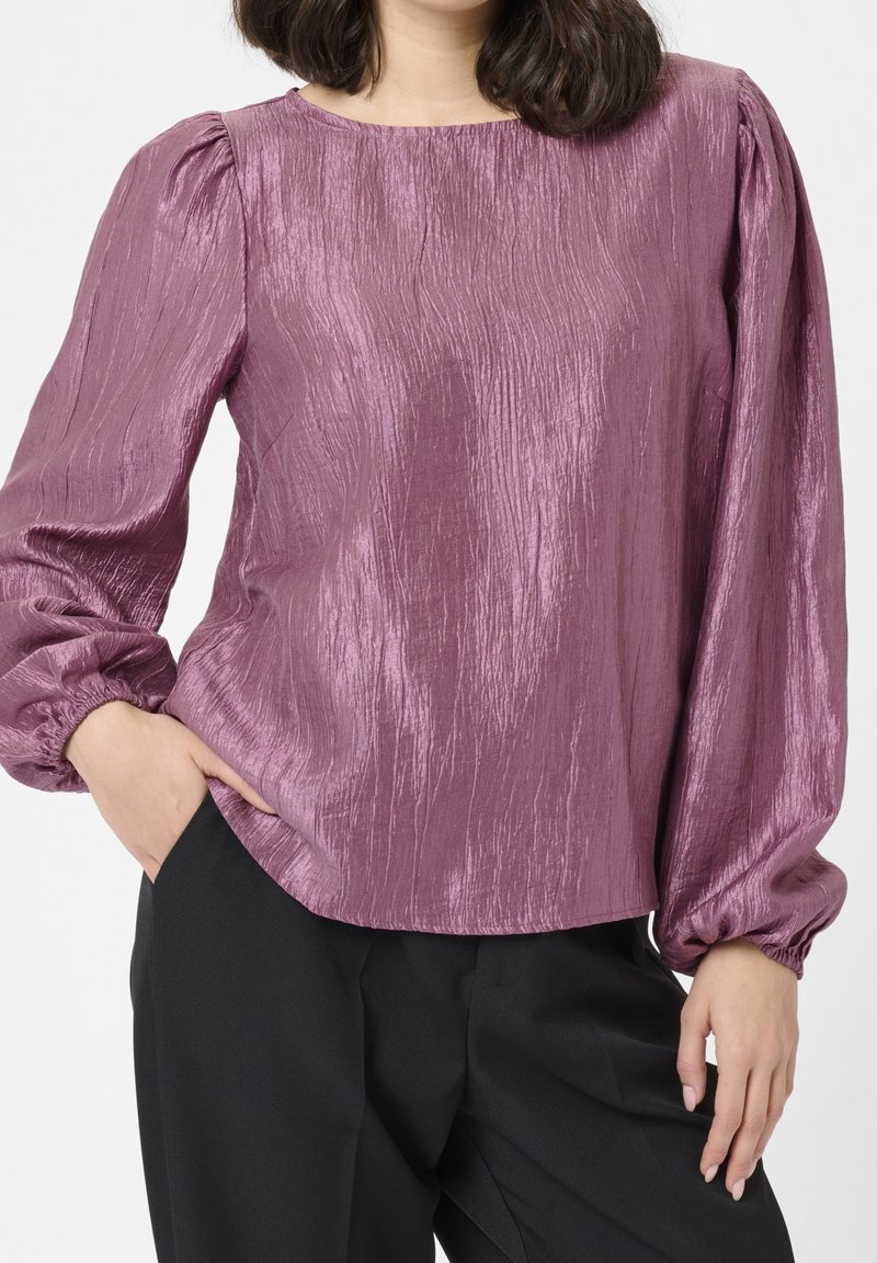 Femme portant un chemisier mauve à manches longues texturé et brillant avec des manches bouffantes, et un pantalon noir, une main dans la poche.
